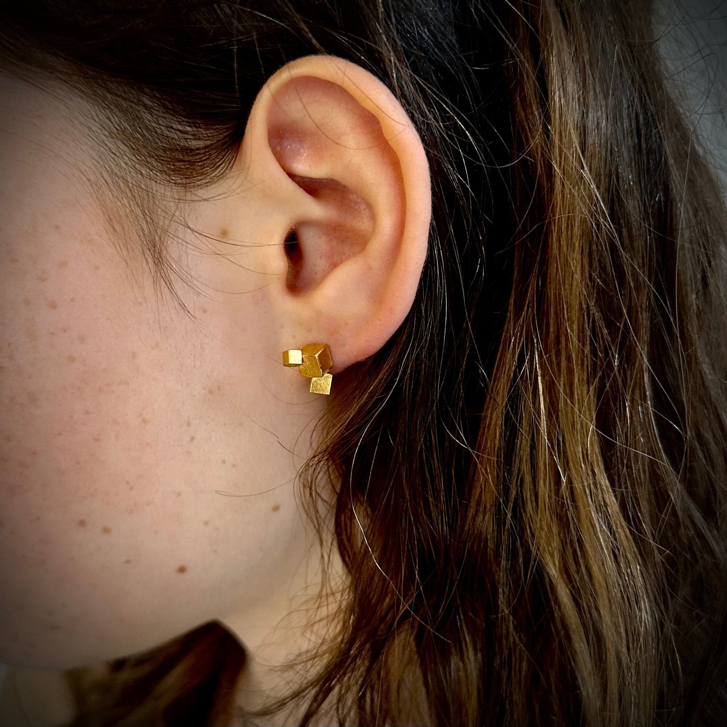 Close-up of an ear wearing gold cubist geometric earrings with dark hair.