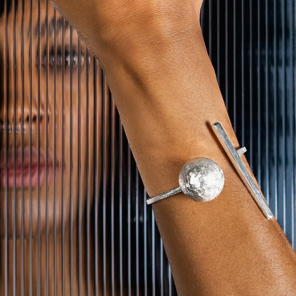 A person wearing a silver sculptural cuff bracelet poses behind a black screen with vertical slits, showing their hands and face through the transparent bars.
