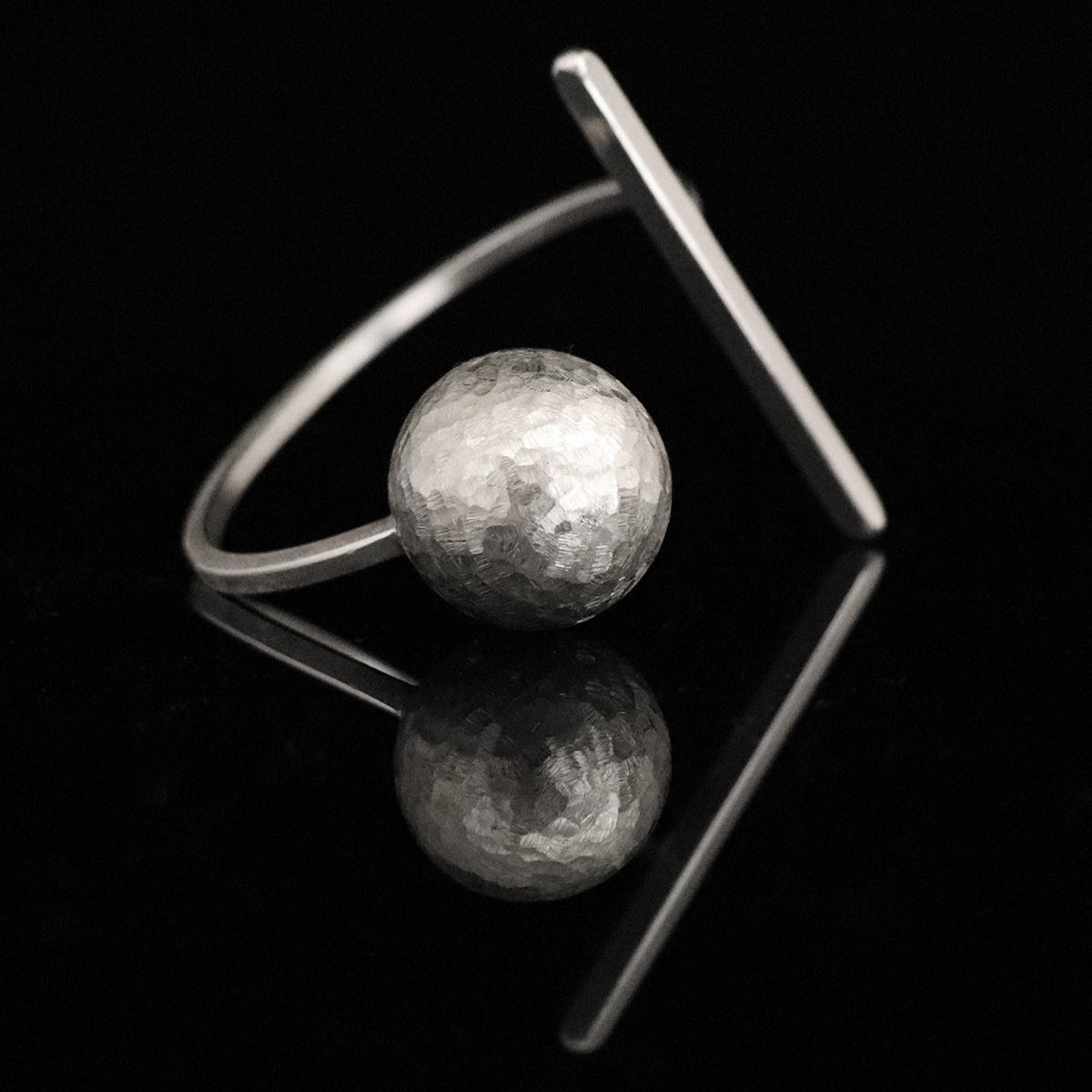 A silver architectural cuff bangle featuring a hammered-texture spherical bead and a flat, angled bar, displayed on a reflective black surface.