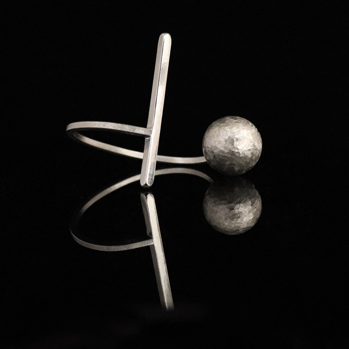 A sculptural silver bracelet cuff featuring a flat, open band with a vertical bar and a textured spherical bead, displayed on a reflective black surface.