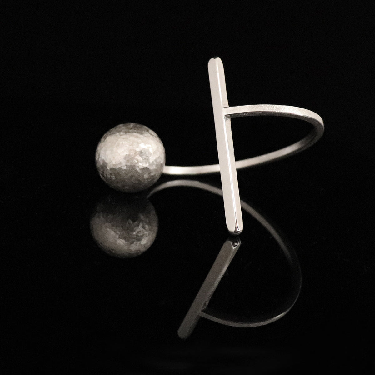A architectural silverbangle cuff featuring a hammered spherical bead and a flat bar, displayed on a glossy black reflective surface.