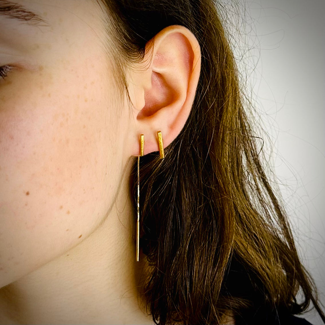 Close-up of an ear wearing gold threader earring and a bar stud with a neutral background