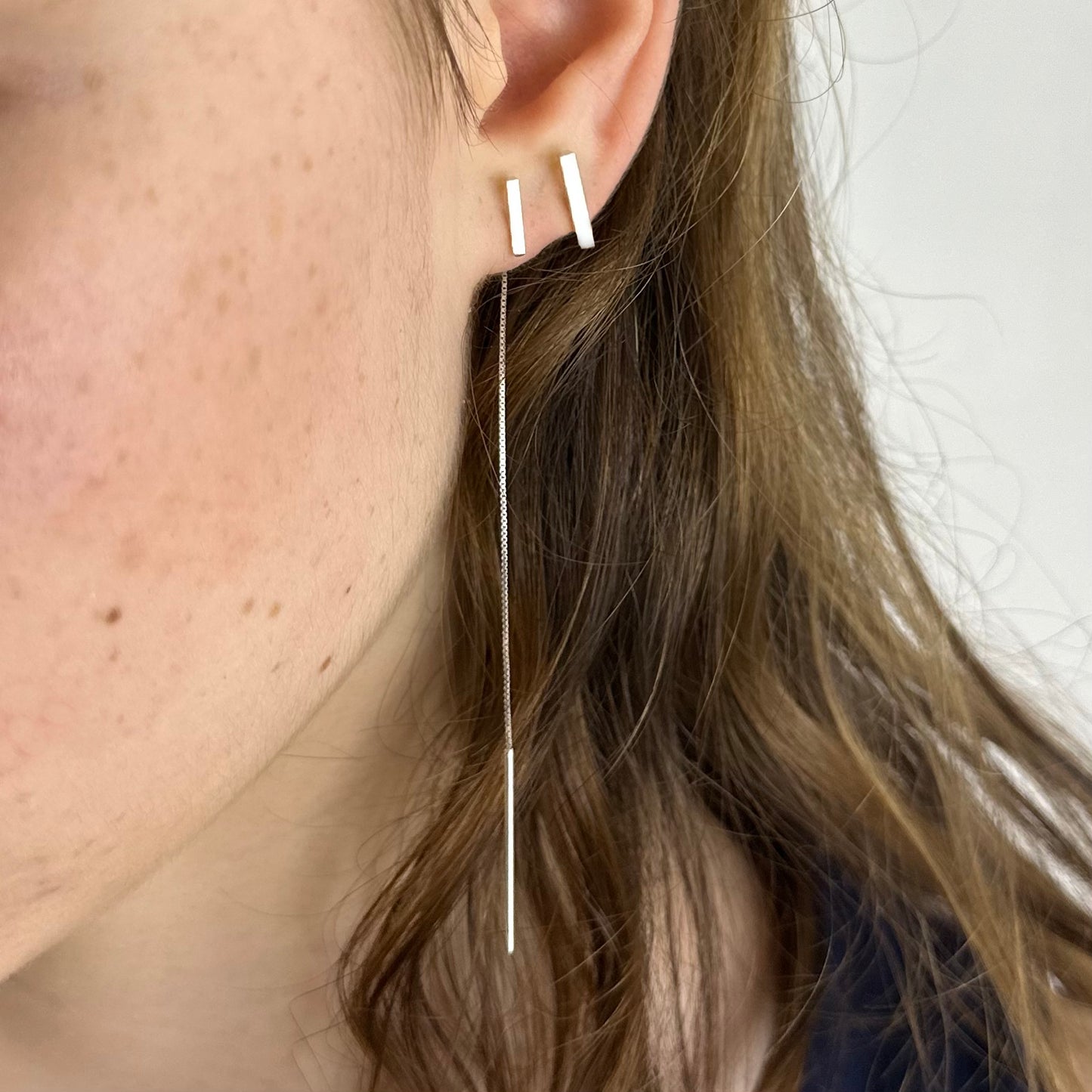 Close-up of an ear wearing a thin silver threader bar earring and a bar stud with a blurred background
