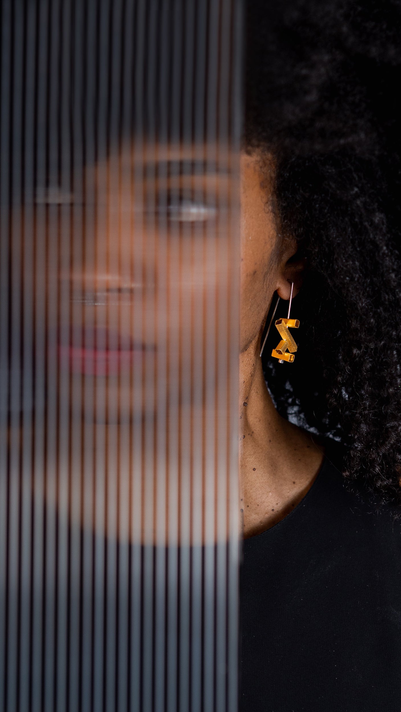 A close-up lifestyle shot of a woman with curly black hair wearing a pair of gold geometric dangle earrings, standing behind a translucent vertical-striped glass panel.