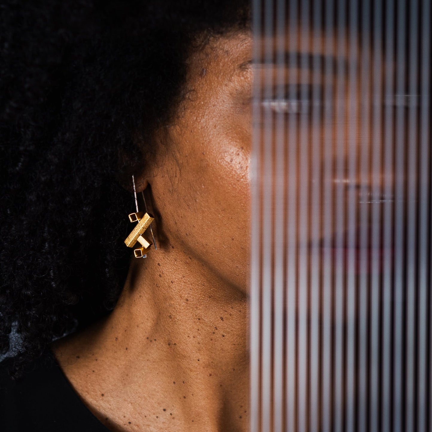 A close-up lifestyle shot of a woman with dark curly hair wearing a gold geometric dangle earring, with a vertical slatted screen partially obscuring her face.