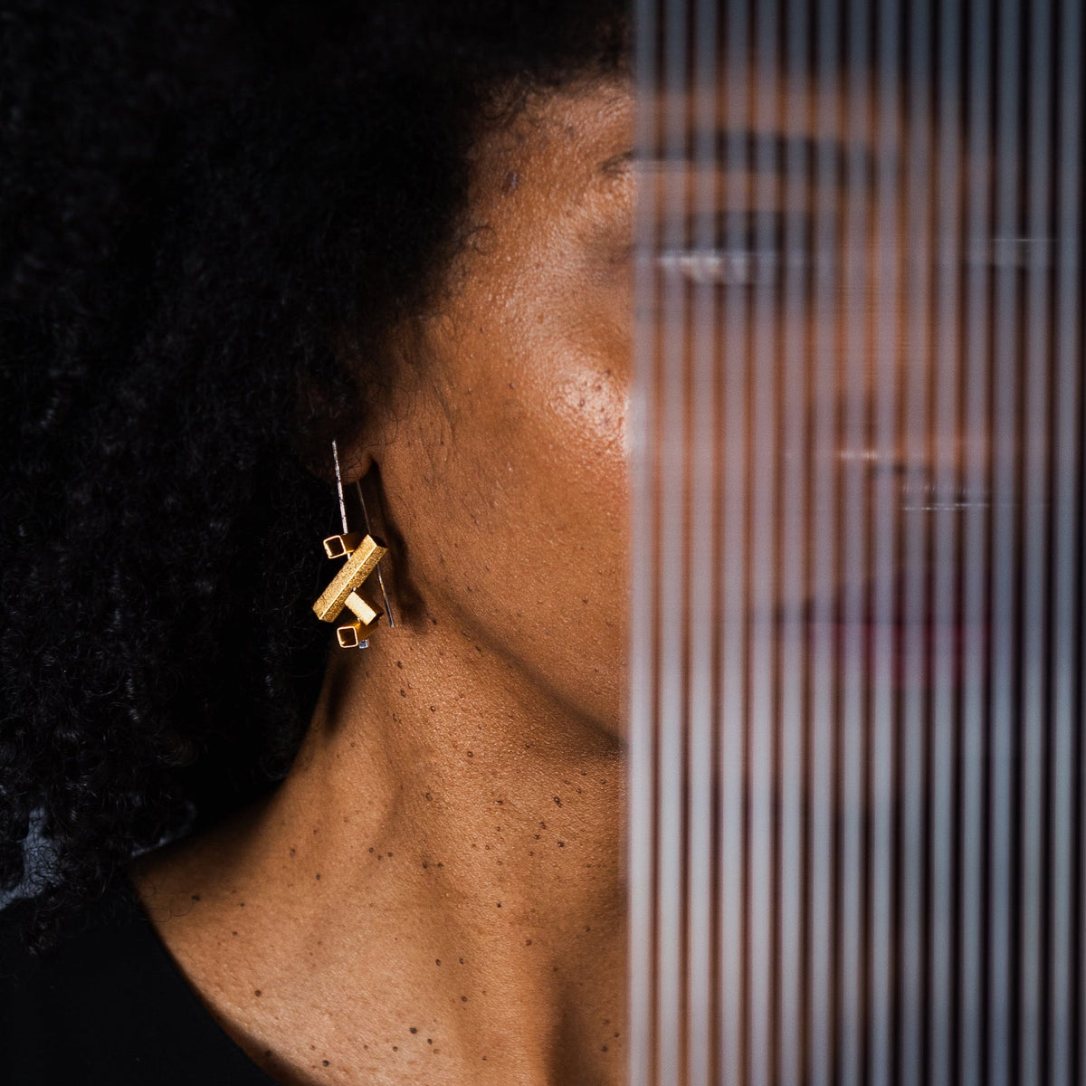 A close-up lifestyle shot of a woman with dark curly hair wearing a gold geometric dangle earring, with a vertical slatted screen partially obscuring her face.