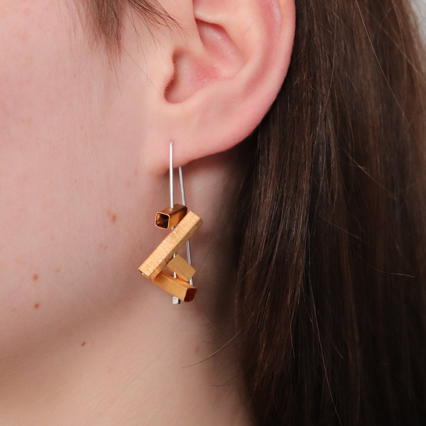 Gold geometric earring with silver wire worn by a person with brown hair.