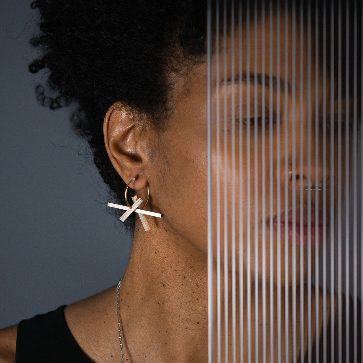 A close-up lifestyle shot of a model wearing a gold geometric cross earring with a small gold hoop, set against a dark gray background with a vertical striped overlay.