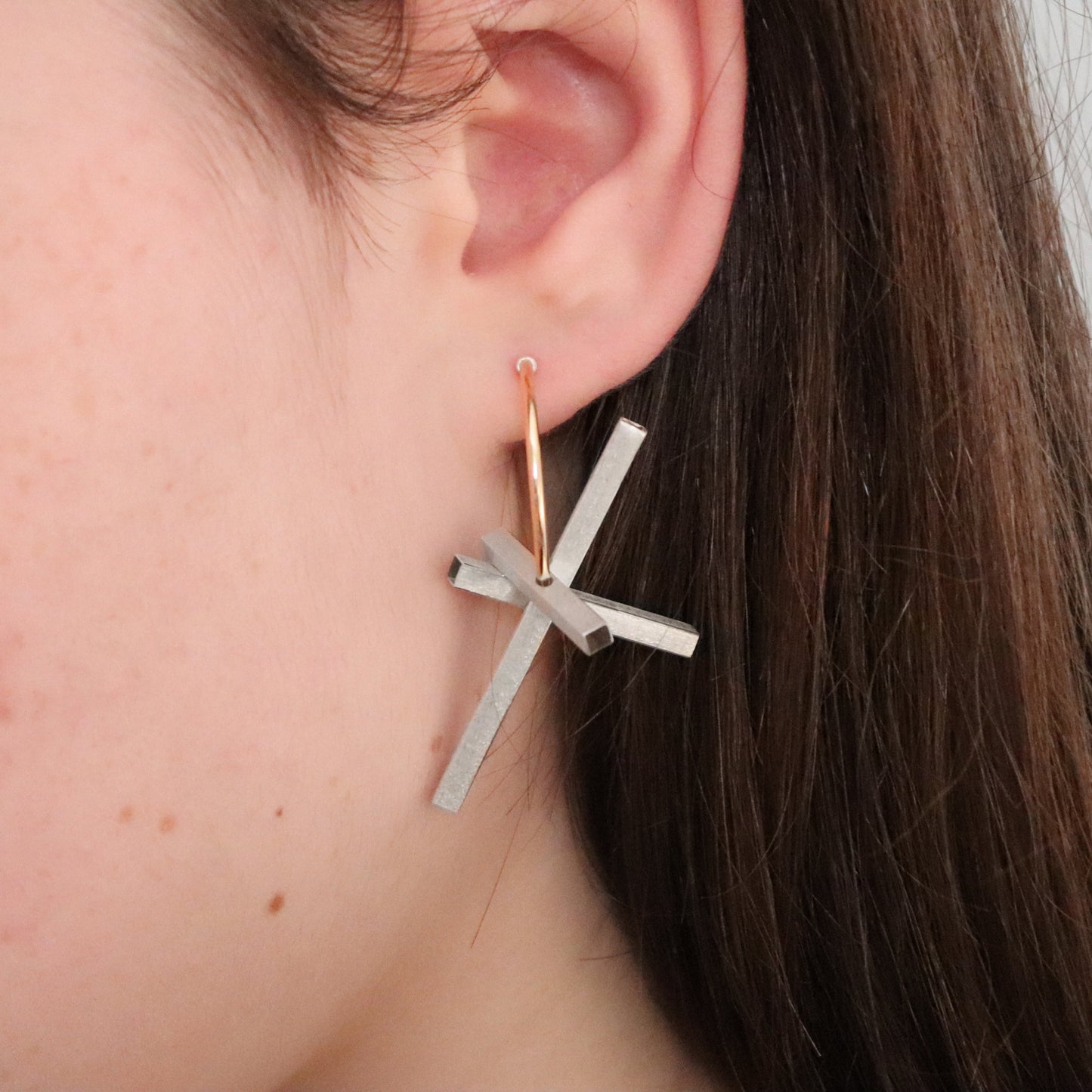 Silver cross-shaped earring worn by a person with brown hair.