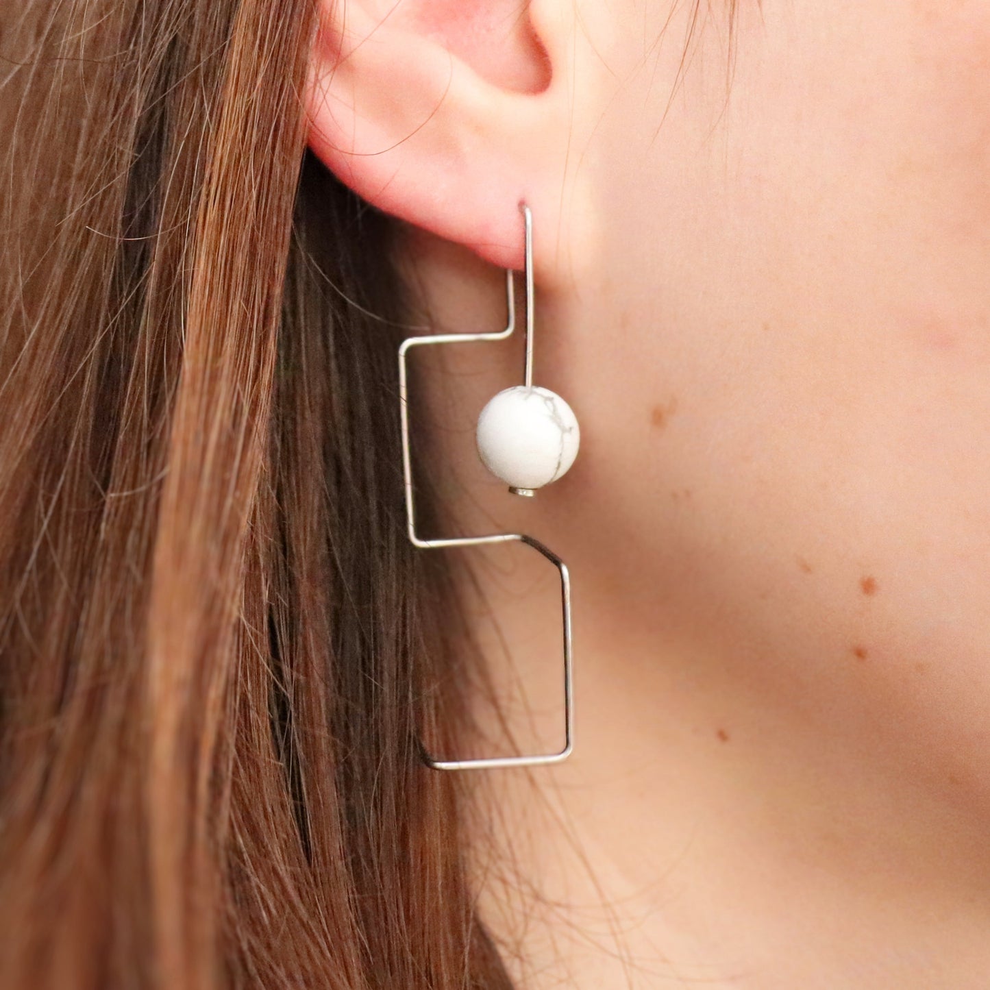 Earring with a spherical howlite bead and geometric frame worn by a person.