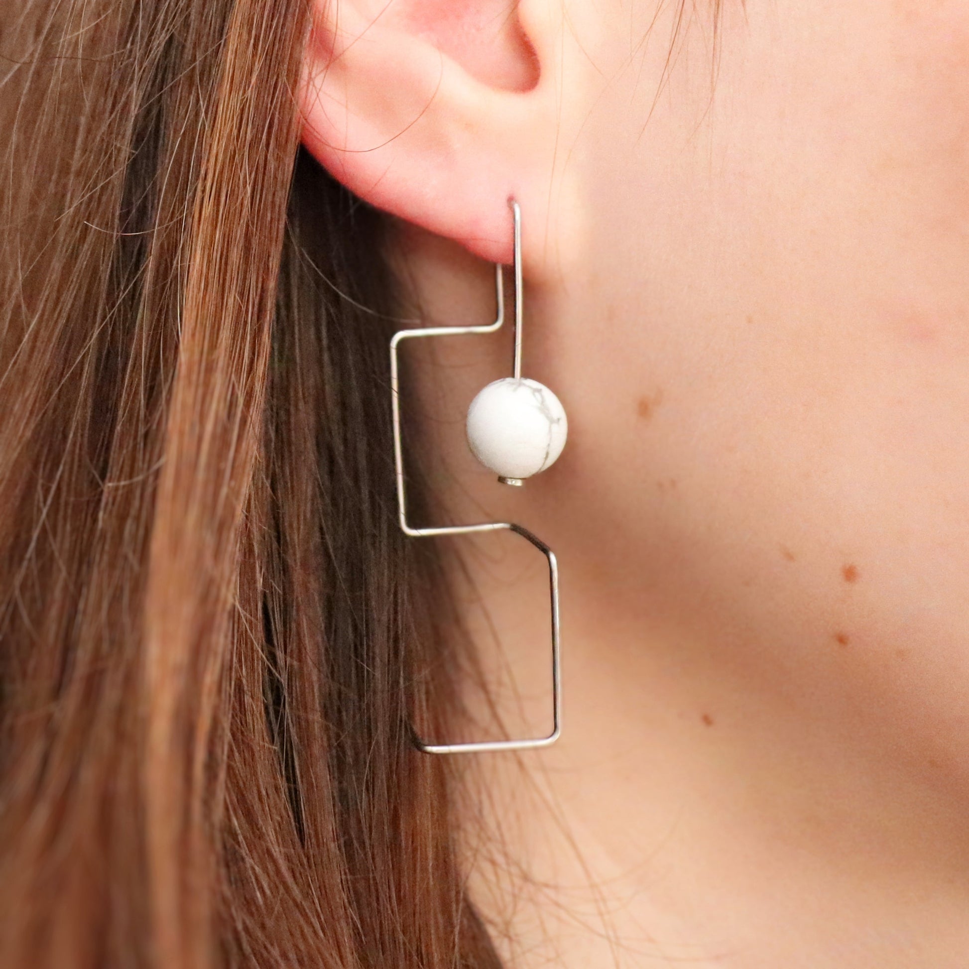 Earring with a spherical howlite bead and geometric frame worn by a person.