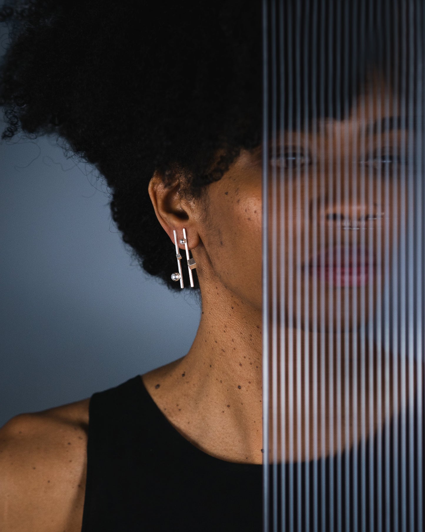 A close-up portrait of a woman with dark curly hair wearing a pair of geometric  silver minimal earrings, set against a dark gray background with a vertical ribbed translucent screen overlay.