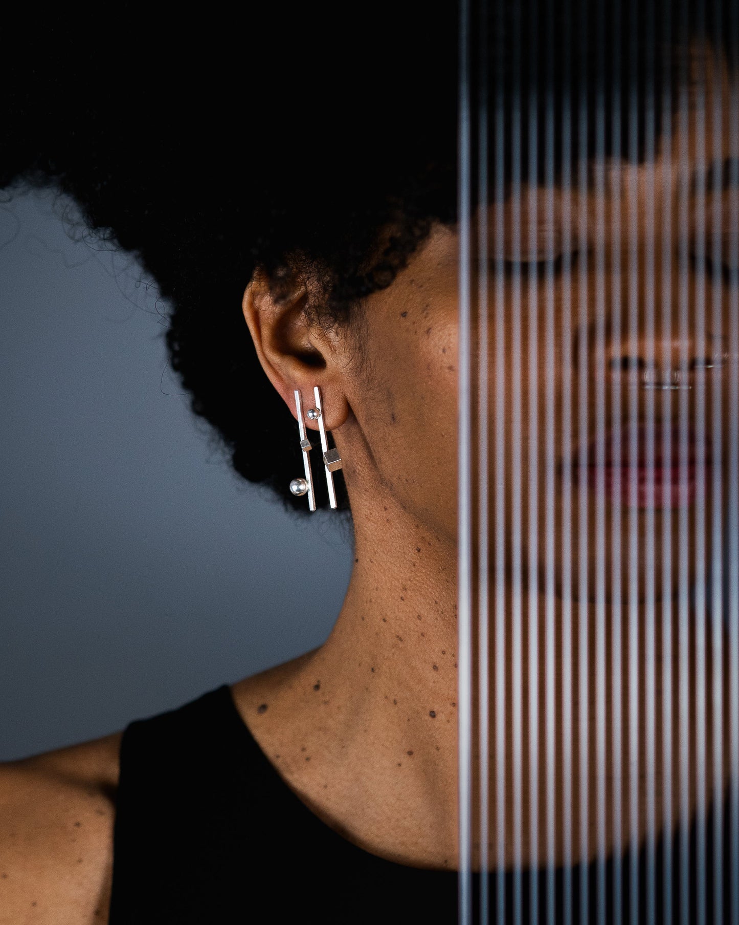 A close-up side profile of a person wearing a pair of silver geometric drop earrings, with a vertical split-screen effect showing a mirrored reflection of the face and neck against a dark gray background.