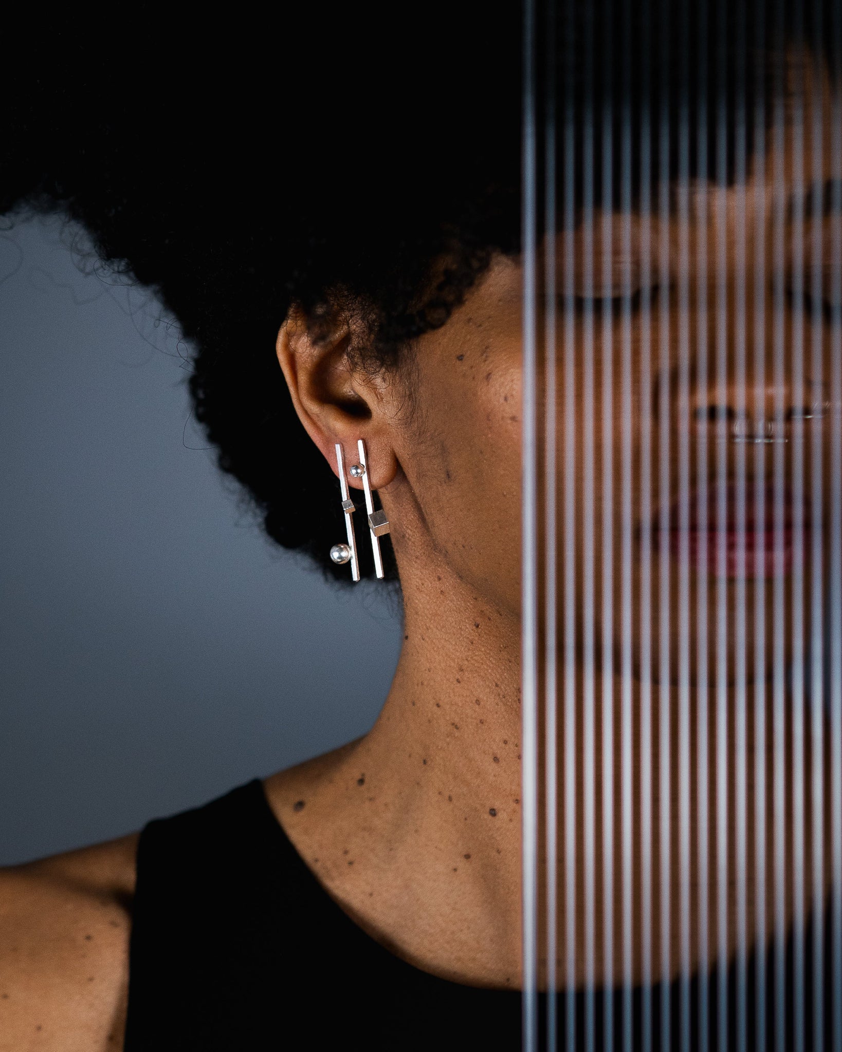 A close-up side profile of a person wearing a pair of silver geometric drop earrings, with a vertical split-screen effect showing a mirrored reflection of the face and neck against a dark gray background.
