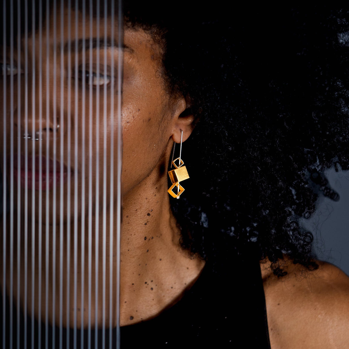 A close-up lifestyle shot of a model wearing a pair of geometric gold earrings with silver hooks, set against a dark background with a vertical slatted glass panel on the left.