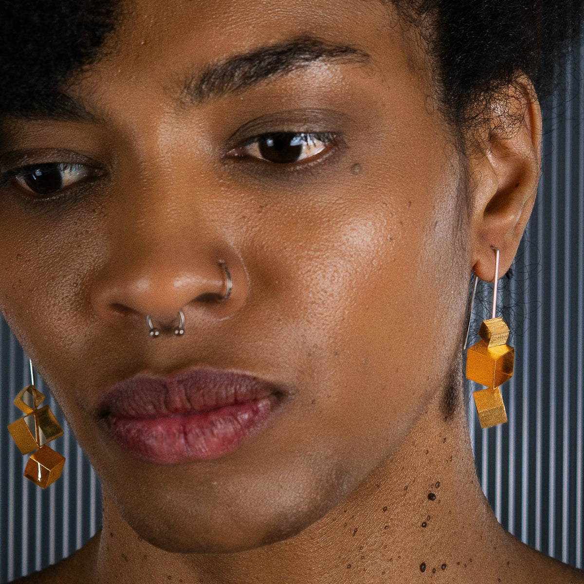 Close-up of a person wearing geometric gold brutalist style earrings with cube-like shapes, shown against a dark striped background.