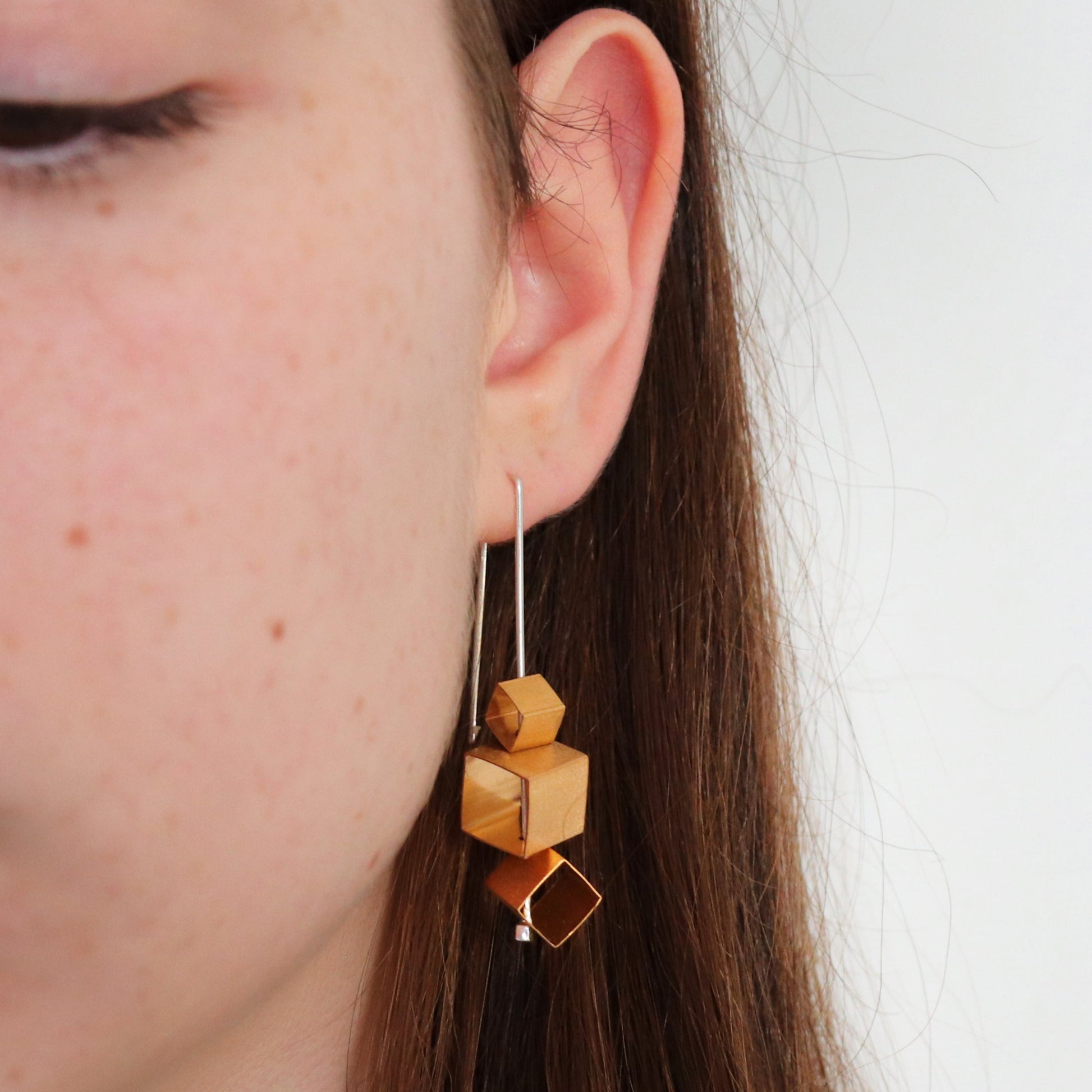 Close-up of a person wearing geometric gold cubes on silver wire earrings with a plain background