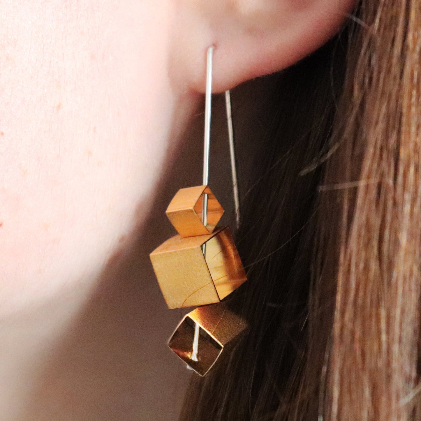 Gold cube-shaped earrings worn by a person with brown hair.
