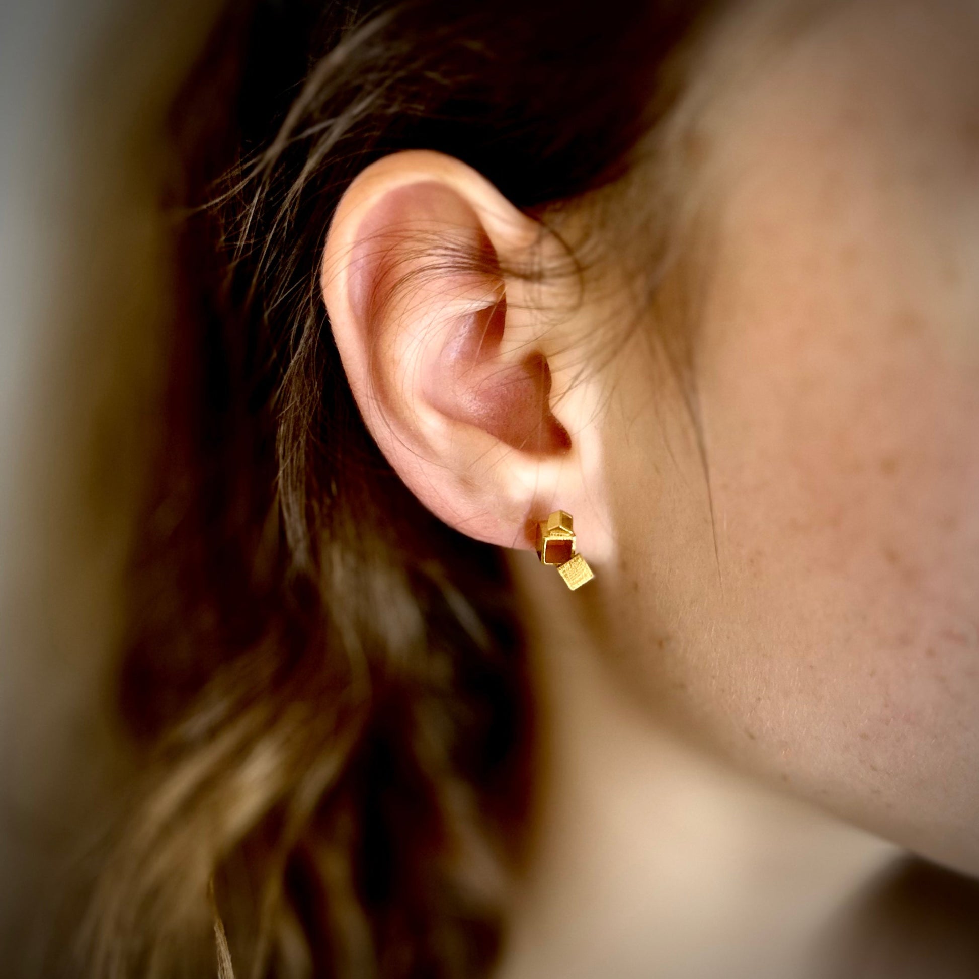 Close-up of an ear wearing a gold architectural cube earring with blurred hair and skin in the background