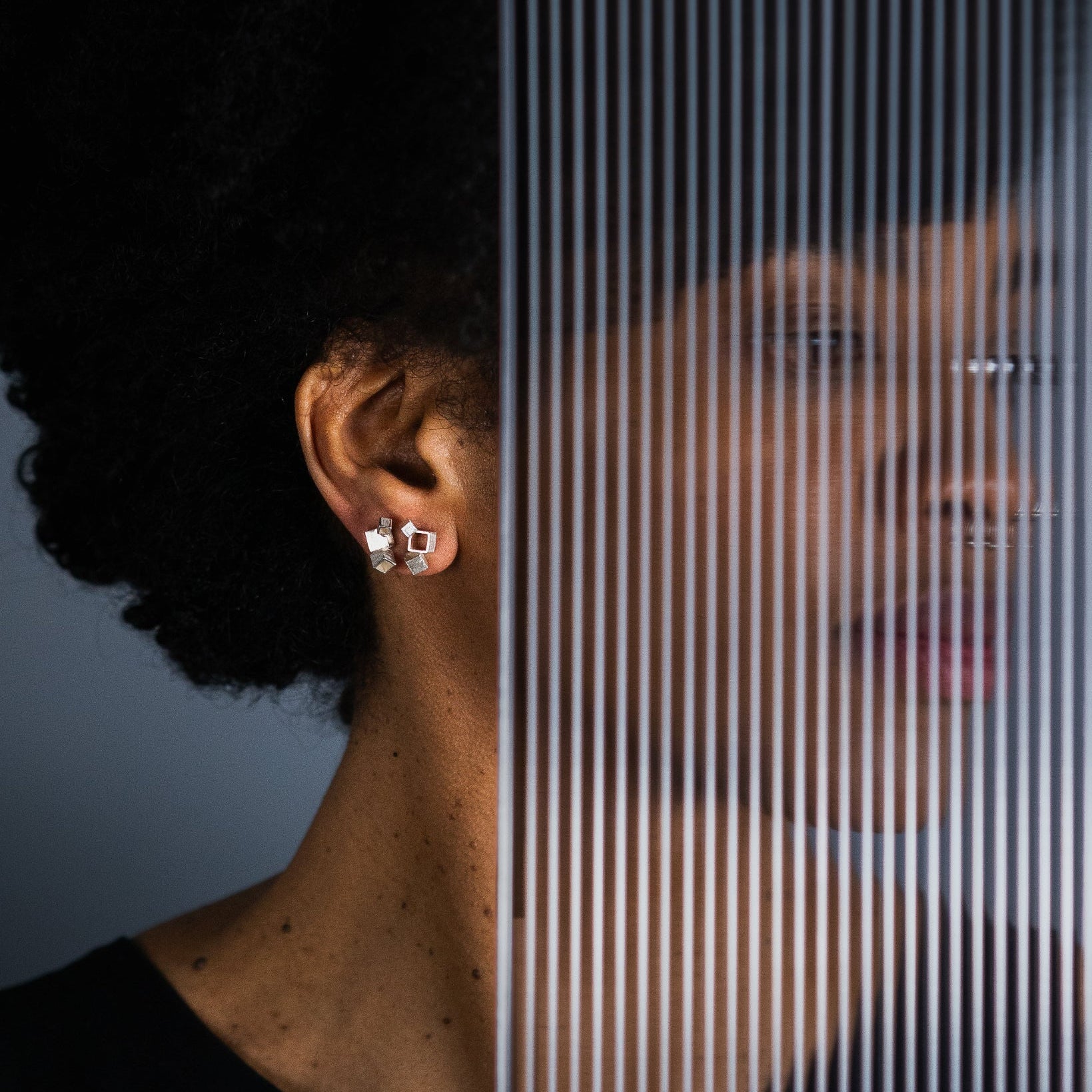 A close-up  of a person with dark curly hair wearing a architectural cube silver stud earring, with a vertical striped translucent screen overlaying the right side of the frame.