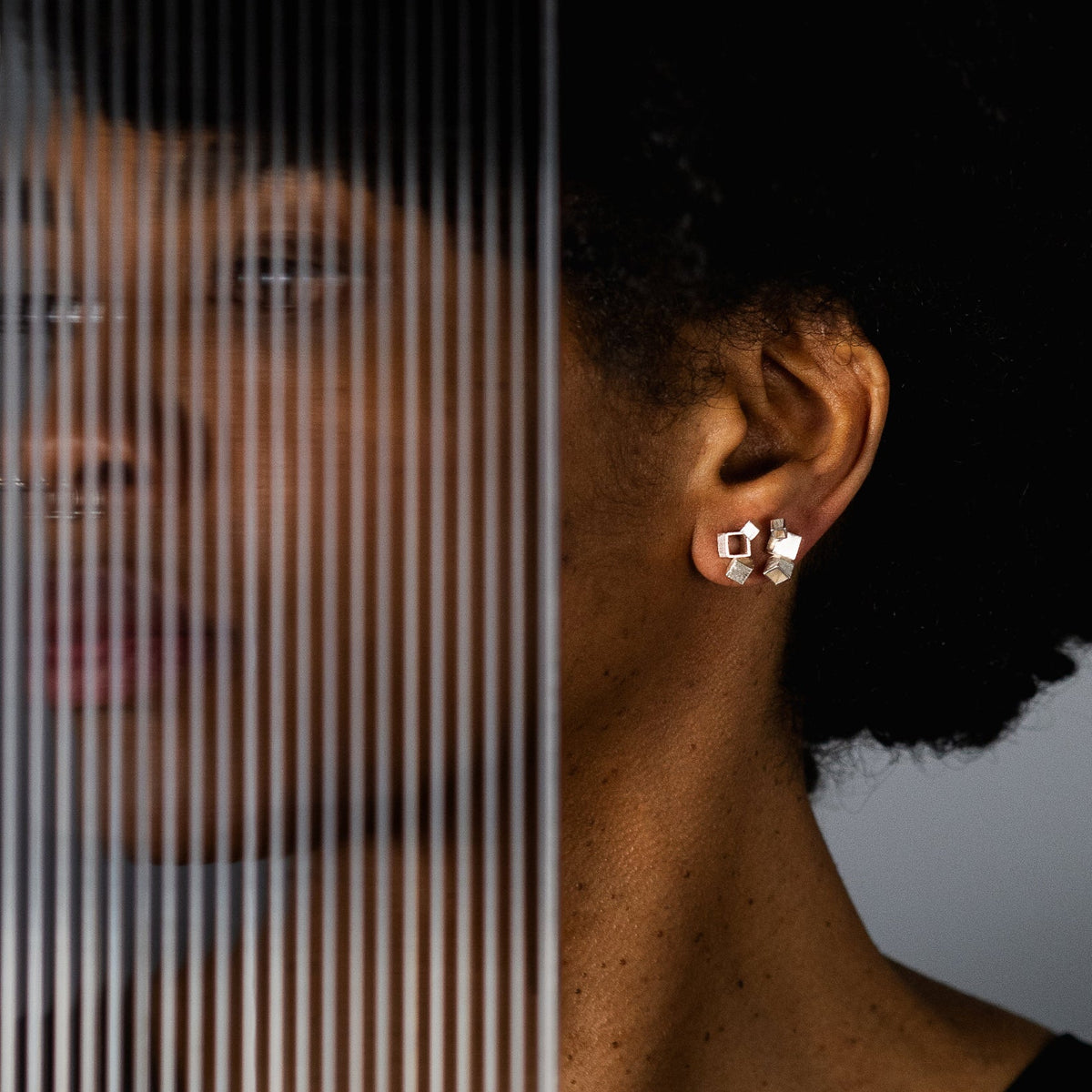 A close-up lifestyle shot of a model's ear wearing a pair of gold geometric stud earrings with clear stones, set against a dark background with a vertical striped glass panel overlay.