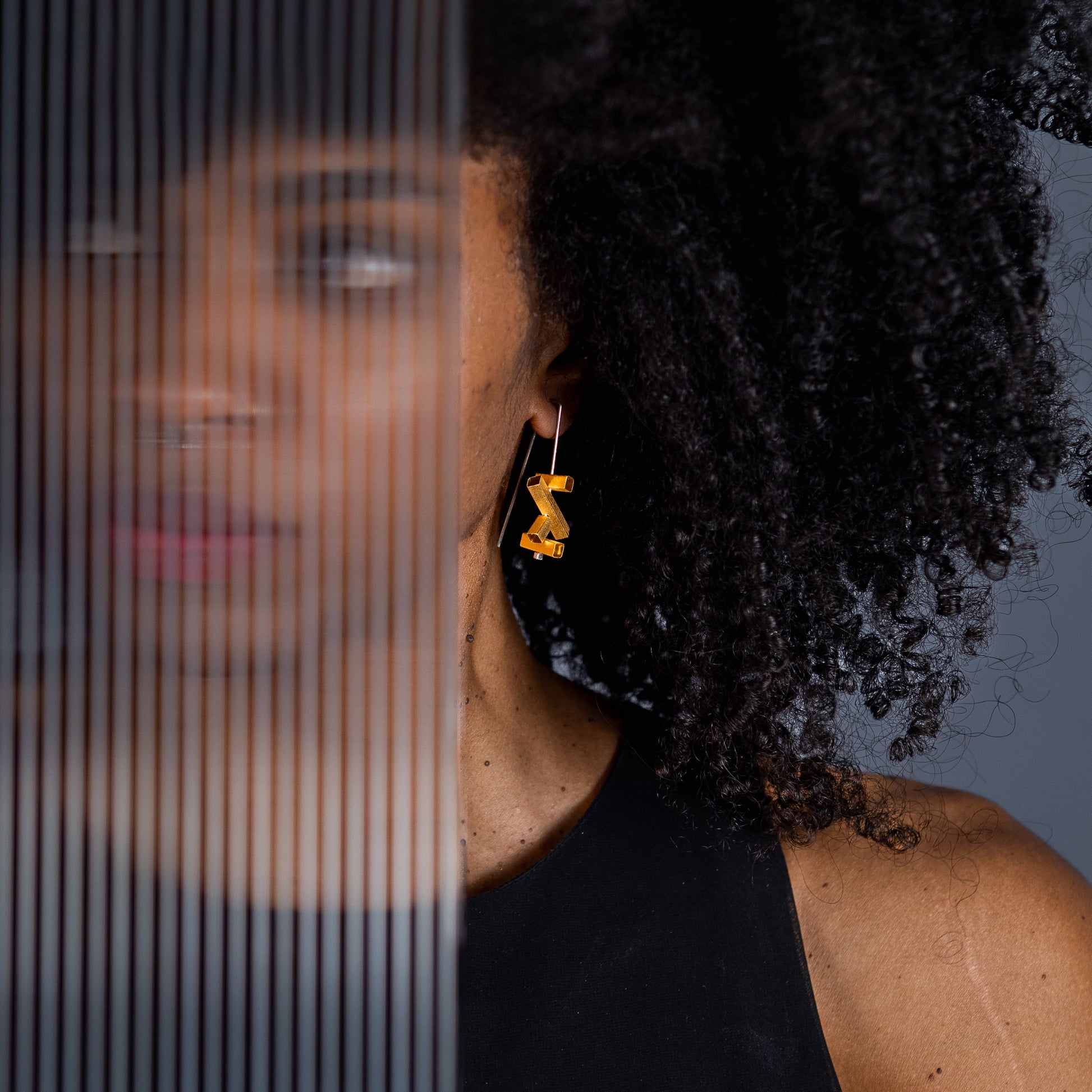 A close-up lifestyle shot of a woman with curly black hair wearing a pair of gold geometric dangle earrings, standing behind a translucent vertical-striped glass panel.
