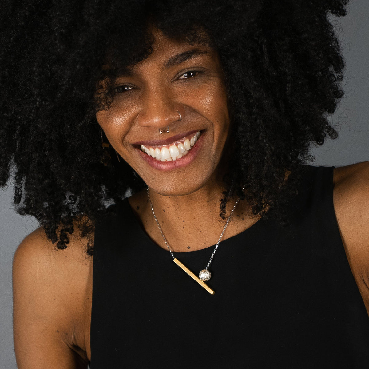 A smiling woman with dark curly hair wearing a minimalistic necklace with a gold bar and silver ball pendant, photographed against a solid grey background.