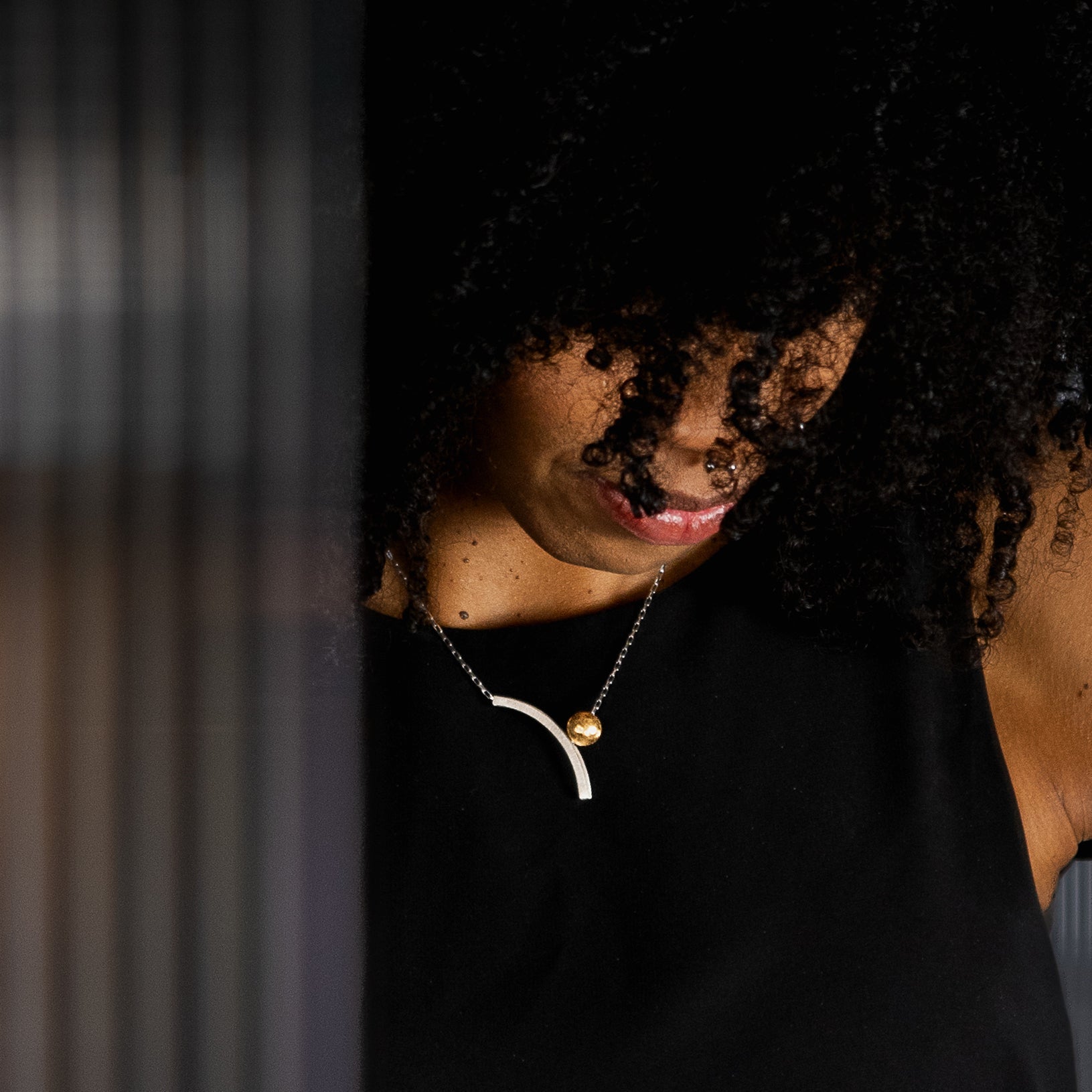 A lifestyle shot of a person with curly black hair wearing a architectural minimal silver & gold necklace, set against a dark, moody background.
