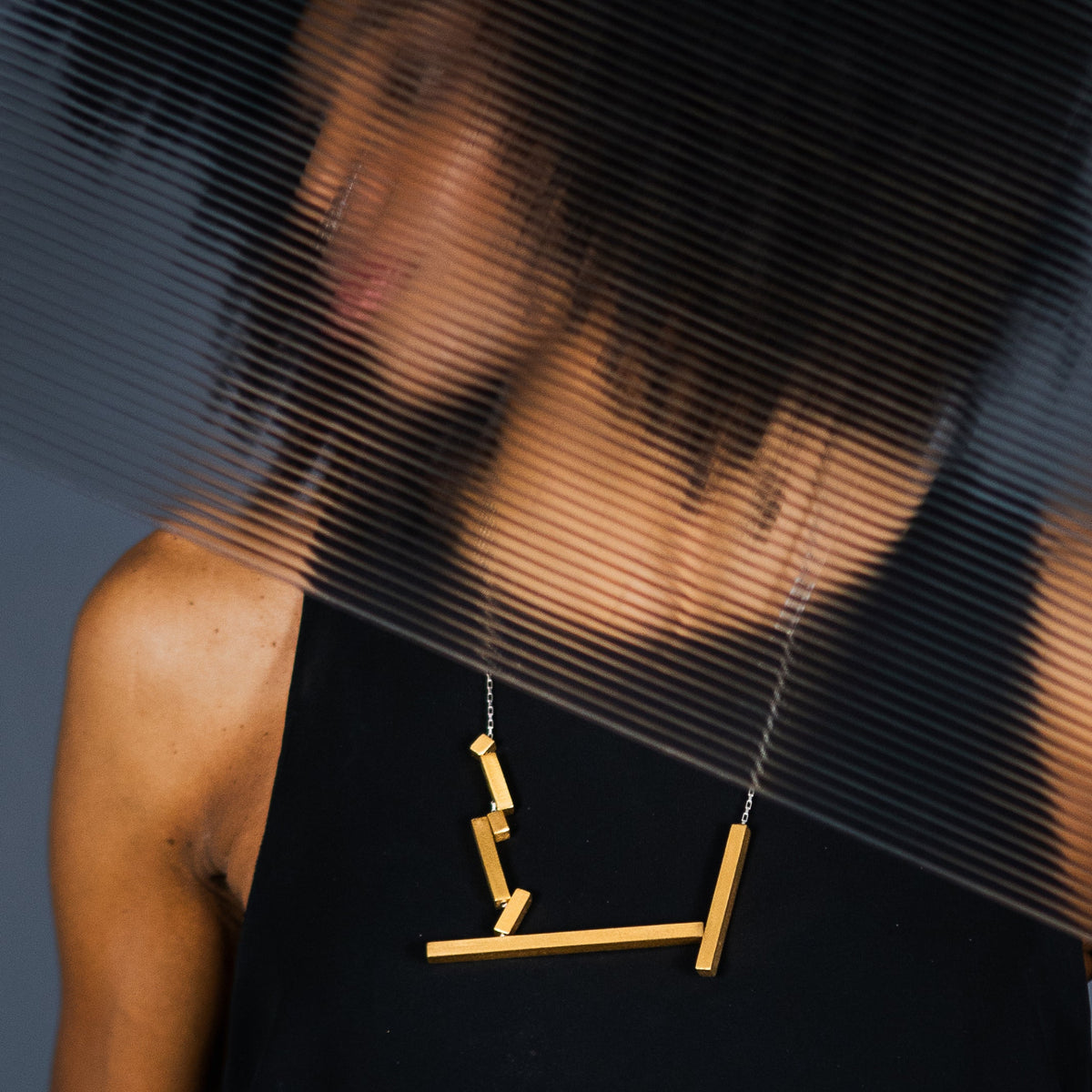 A person wearing a black sleeveless top and a gold geometric pendant necklace, photographed with a translucent ribbed overlay across the upper frame.