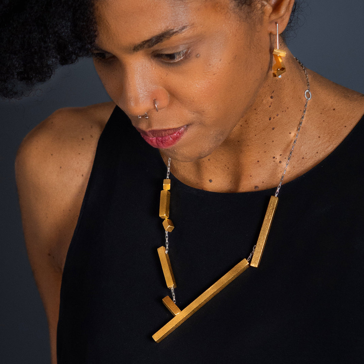 A close-up of a woman wearing a modern statement geometric gold necklace and matching gold earrings earrings, styled with a black sleeveless top against a dark gray background.
