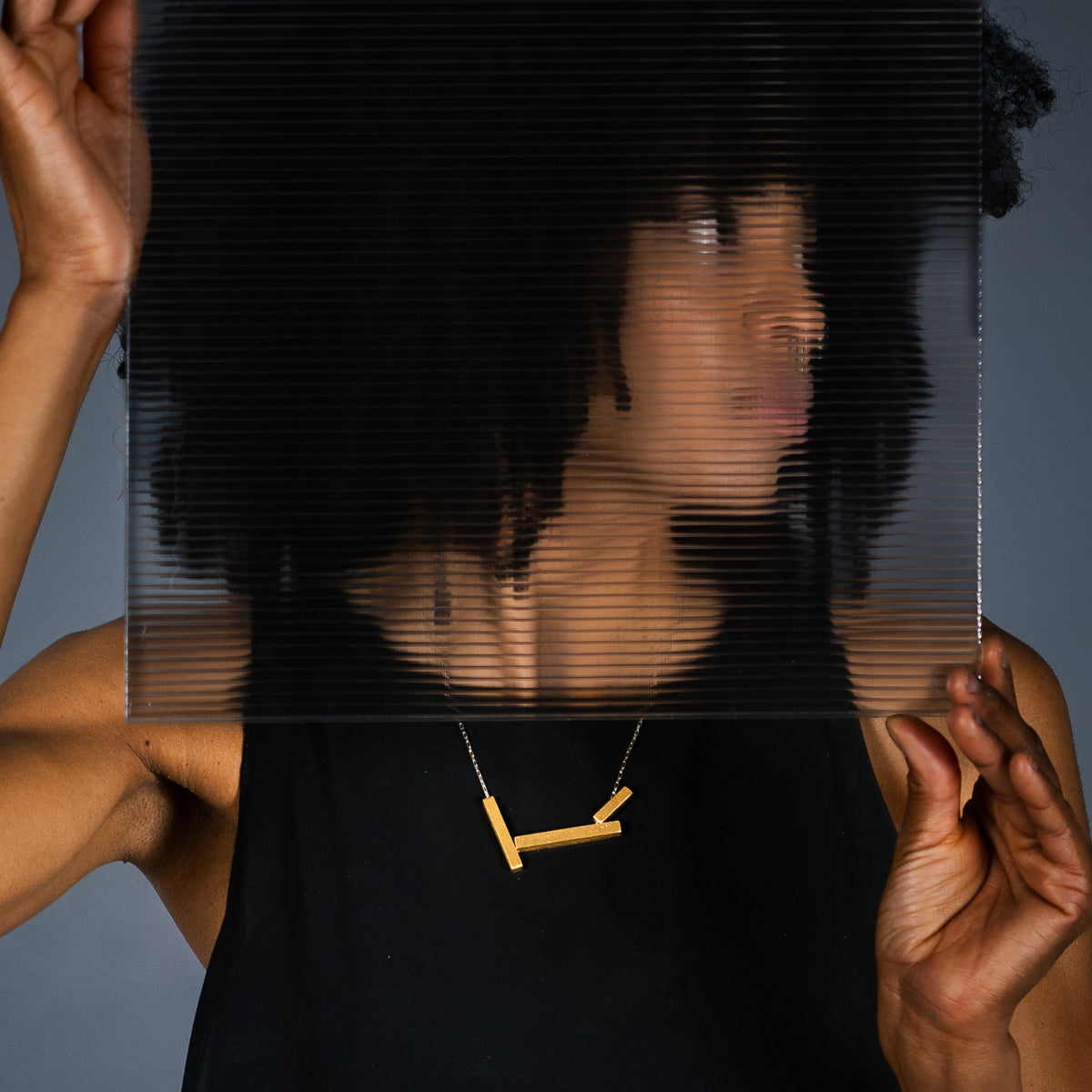 A model with dark curly hair wearing a gold geometric statement necklace, holding a translucent black ribbed square panel in front of their face against a dark gray background.