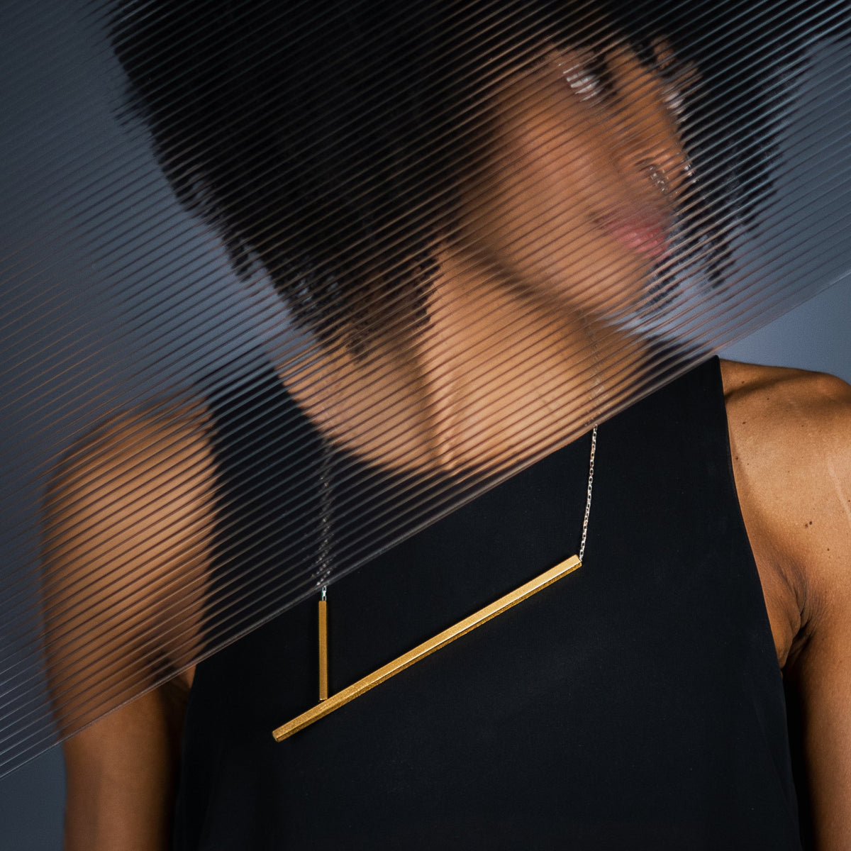 A woman wearing a minimalist gold geometric minimal statement necklace with a horizontal bar and a vertical drop, styled with a black sleeveless top against a dark grey background with a diagonal striped overlay.