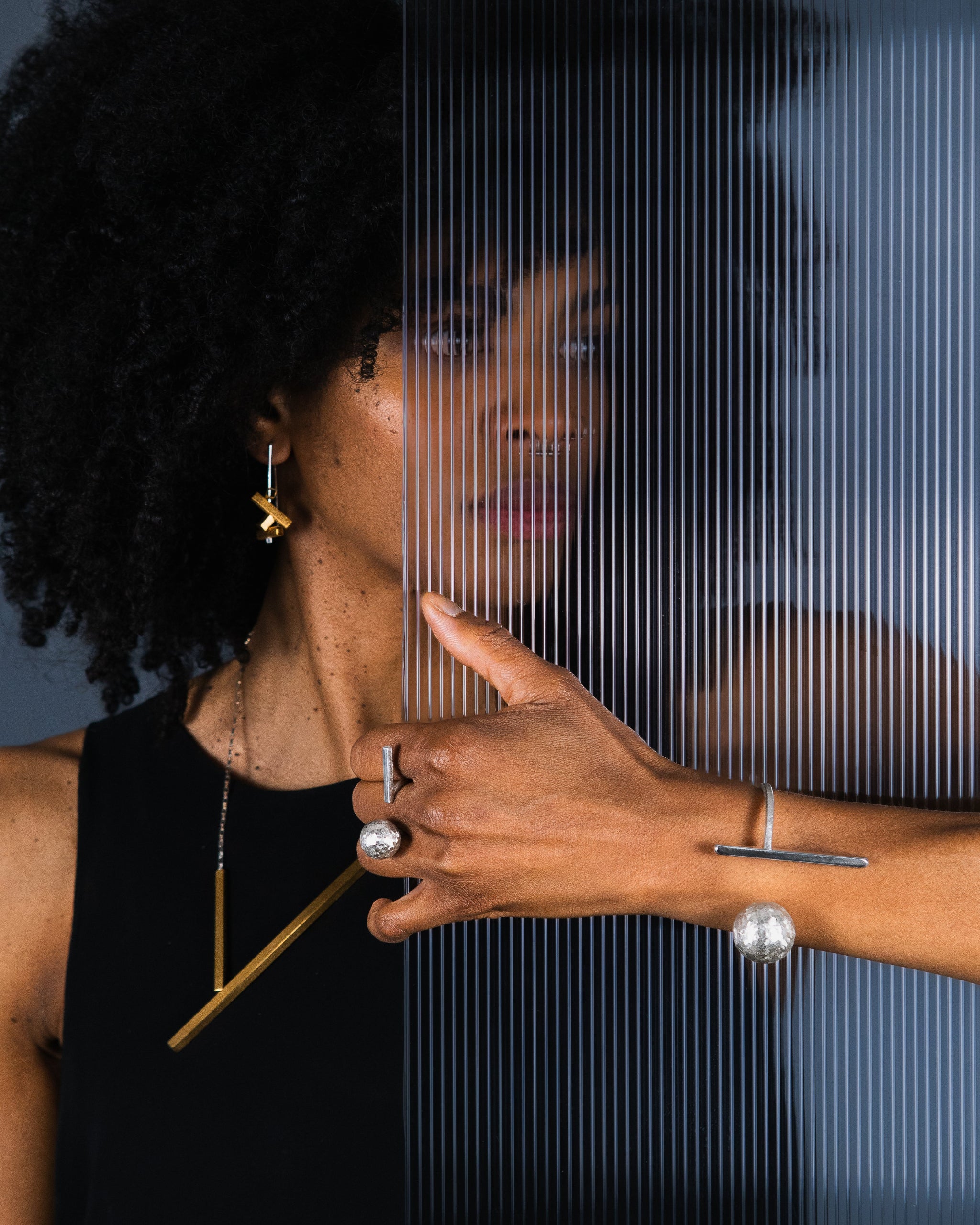 A close-up lifestyle shot of a model with dark curly hair wearing minimalist geometric jewelry, including a gold earring, a gold necklace, a silver ring, and a silver bracelet, posed behind a glass pane with vertical lines.