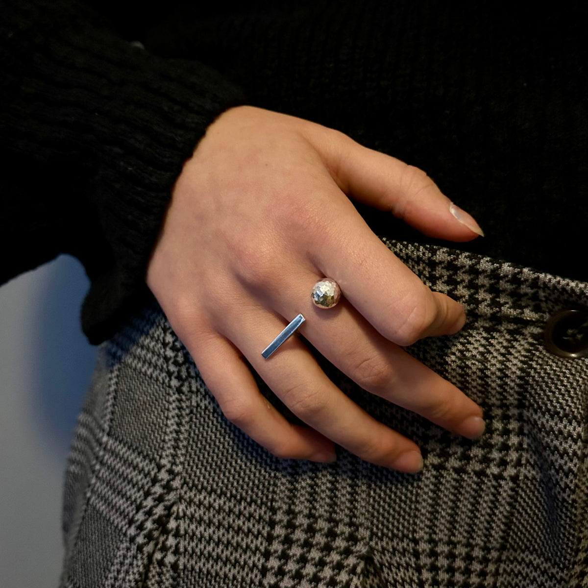 A close-up of a person's hand wearing two minimalist silver rings, one with a small round silver ball and the other with a flat bar, resting on a black and white houndstooth skirt.