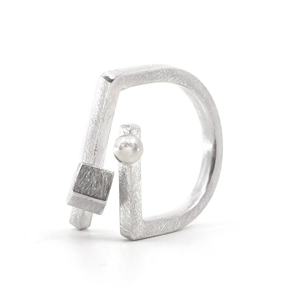 A silver ring featuring an abstract, open geometric design with a textured finish, a small square accent, and a ball, shown on a plain white background.