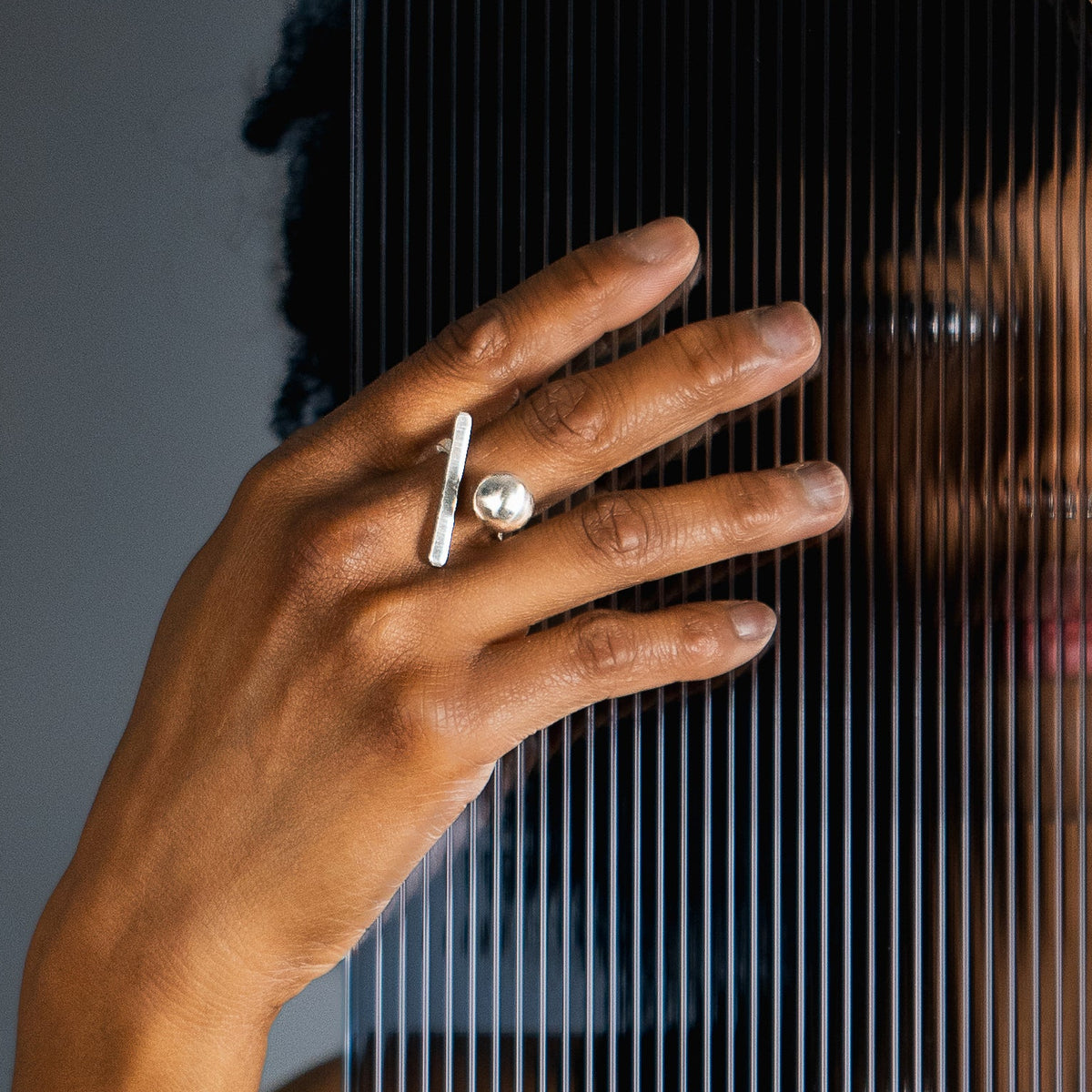 A person wearing a silver cocktail ring with a ball and bar geometric component poses behind a black screen with vertical slits, showing their hands and face through the transparent bars.