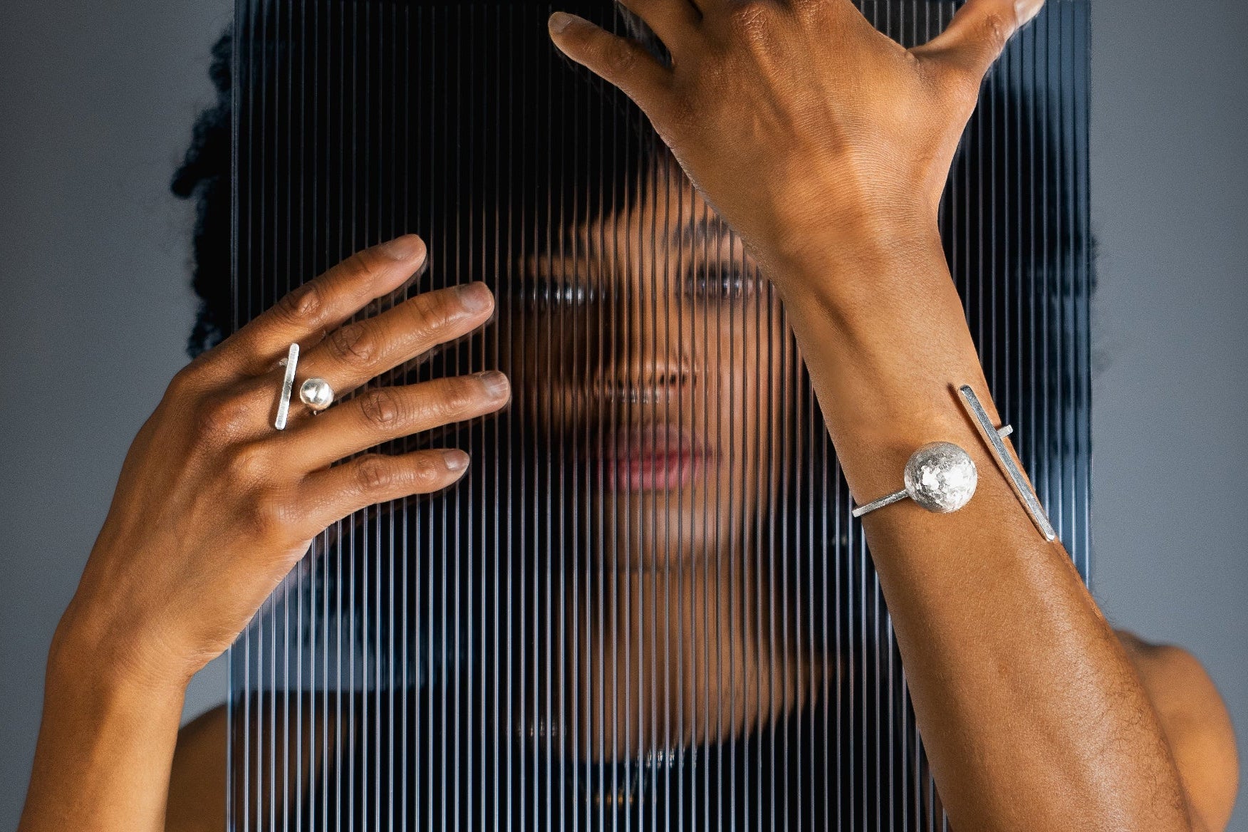 A close-up lifestyle shot of a person wearing silver jewelry, including a ring on one finger and a cuff bracelet on the wrist, with a blurred face visible behind vertical bars.