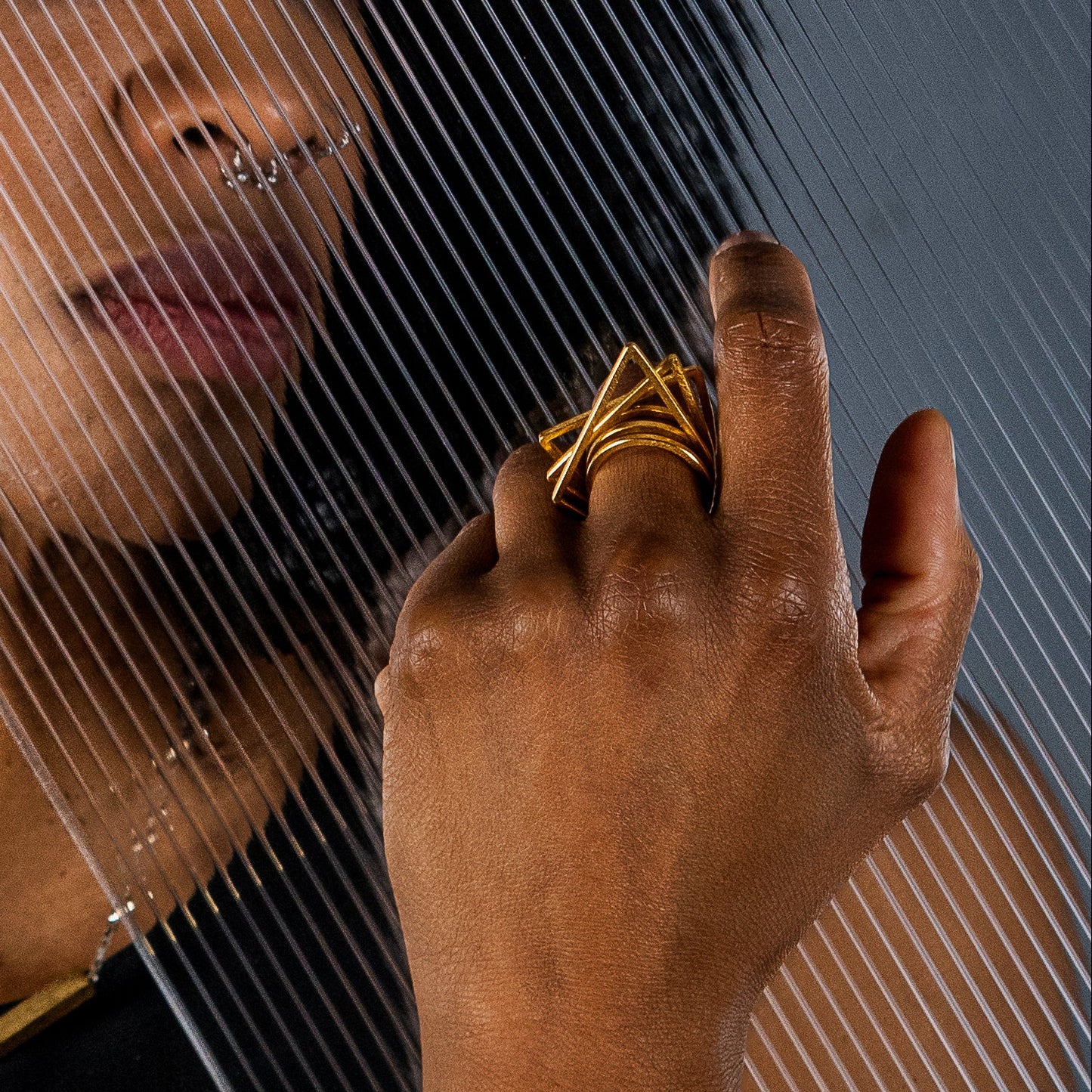 A close-up portrait of a model with dark curly hair wearing gold geometric stacking rings, behind a translucent diagonal panel.